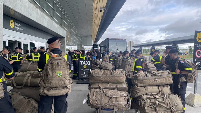 Des dizaines de pompiers attendent avec leurs bagages de monter dans un autocar sur le quai du stationnement de l'aéroport de Québec.