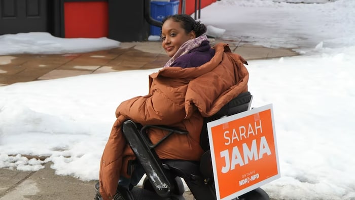 سارة جاما على كرسي متحرك وعلى ظهرها ملصق برتقالي اللون (لون حزبها) يحمل اسمها.