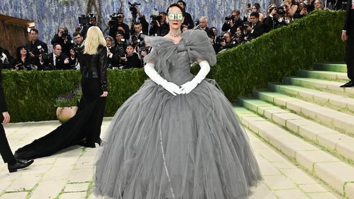 Sarah Paulson porte une robe grise et un billet de banque lui couvre les yeux. 