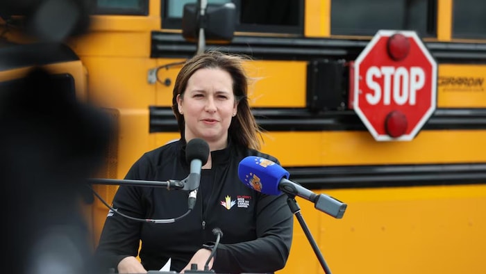 La ministre debout dehors, devant un autobus scolaire.