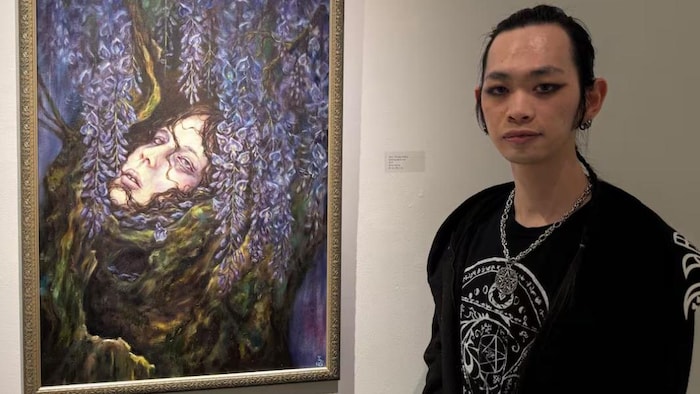  Qiming Sezava Sun pose à côté d'un tableau mélancolique représentant le visage d'une femme niché dans un tronc d'arbre et entouré de fleurs de glycine violettes.