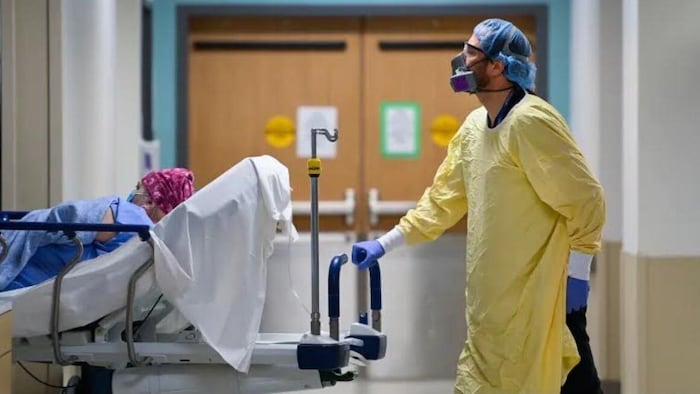 Une personne en combinaison médicale pousse une civière sur laquelle est couchée une autre personne dans un hôpital.
