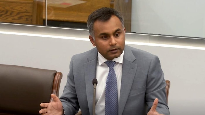 Un homme qui porte un veston gris est assis à un pupitre et est en train de parler.