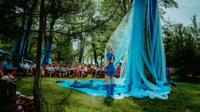 Une femme devant des voiles suspendus à un arbre face à un public assis en forêt.