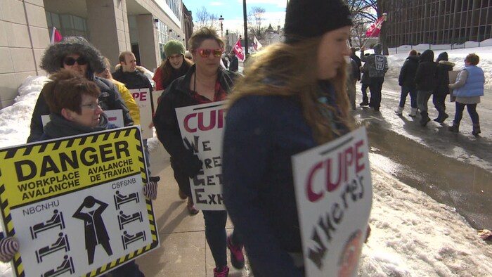 Des gens manifestent sur un trottoir à Fredericton en brandissant des pancartes dénonçant leur surcharge de travail.