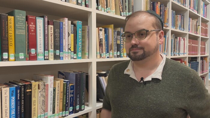 Un homme avec un petit micro épinglé au col de sa chemise est debout devant une bibliothèque remplie de livres.
