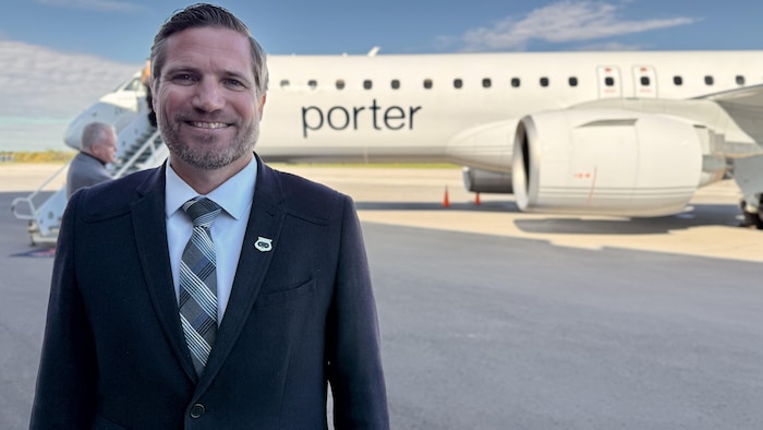 Scott McFadzean sourit devant un avion blanc de Porter Airlines stationné sur le tarmac.