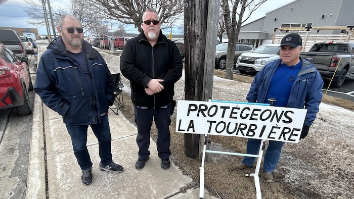 Trois personnes regardent la caméra, une affiche indiquant «Protégeons la tourbière» devant eux.