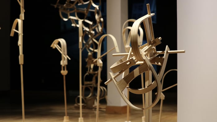 Une sculpture de bois se trouve dans une salle d'exposition, avec plusieurs autres œuvres derrière elle. 