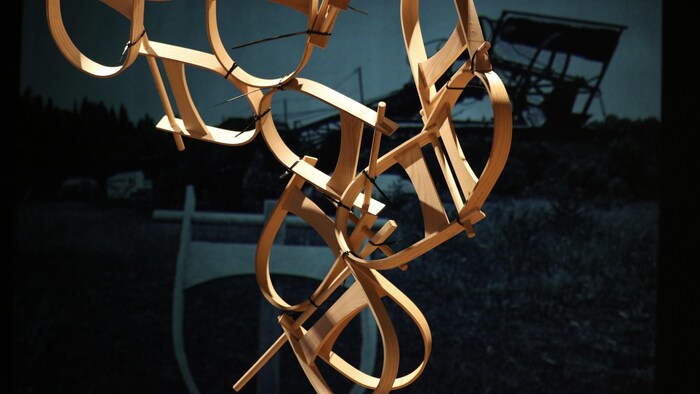 Une sculpture de bois se tient devant une projection sur un mur, dans une salle d'exposition. 