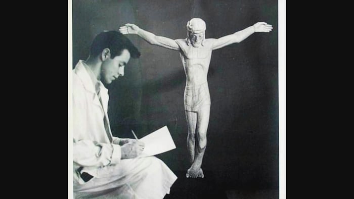 Un jeune Claude Roussel prend des notes à côté de la sculpture.