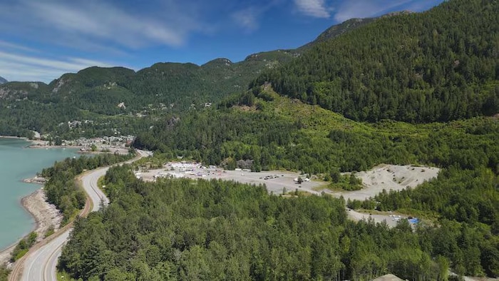 Vue aérienne d'un vaste terrain de gravier aménagé en plateau, bordé d'un côté par une route et une voie ferrée longeant un plan d'eau turquoise, de l'autre par une forêt dense au pied de montagnes; quelques véhicules et bâtiments temporaires occupent le terrain.