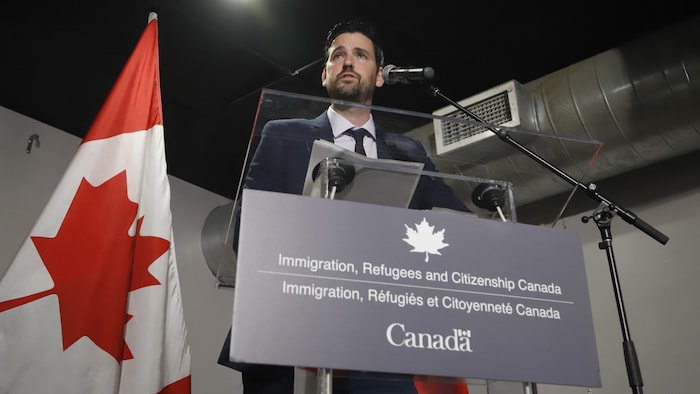 وزير الهجرة واللاجئين والمواطَنة الكندي شون فرايزر يلقي كلمة واقفاً خلف منبر، أمامه ميكروفون وعن يمينه علم كندا.