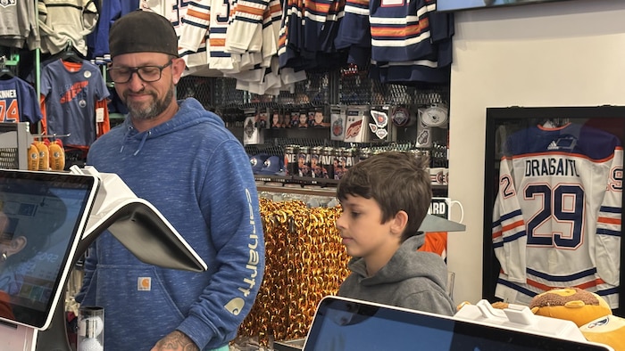 Un homme et un garçon à la caisse d'un magasin, avec des produits des Oilers d'Edmonton en arrière-plan.