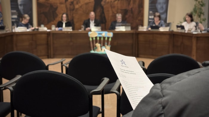 Un citoyen tient l'ordre du jour d'une séance de conseil municipal, assis dans une salle de l'hôtel de ville de Baie-Comeau.