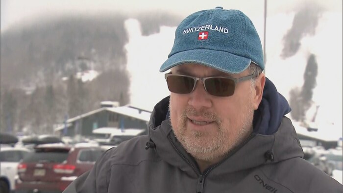 Un homme devant des pistes de ski, avec lunettes fumées et casquette. 