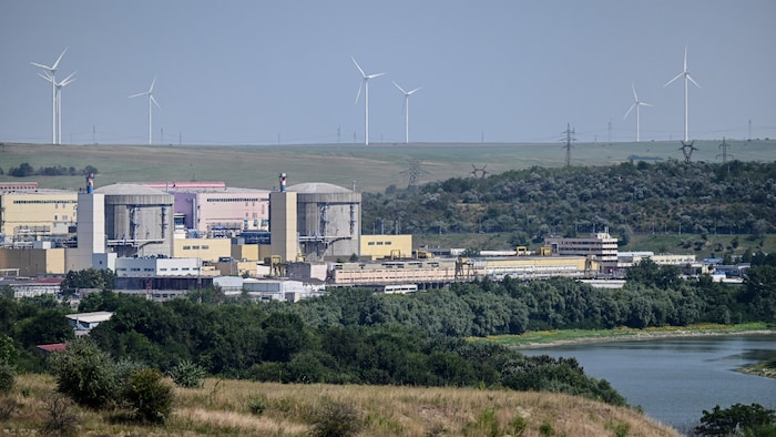 Vue sur une centrale nucléaire au bord d'un cours d'eau, avec à l'arrière-plan des éoliennes.