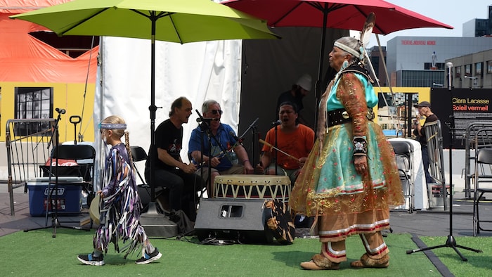 Un enfant et une femme en régalia font une danse traditionnelle devant un groupe de chanteurs qui frappent sur un tambour. 
