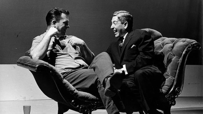 Dans un studio de télévision, l'écrivain américain Jack Kerouac et l'animateur Fernand Seguin, riant, sont assis sur une causeuse.