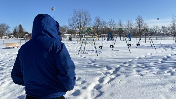 صورة ظهرية لرجل يقف وسط الثلج في حديقة فارغة أمام ملعب للأطفال