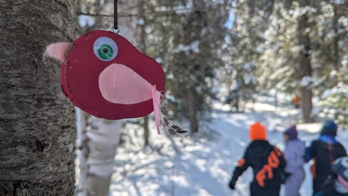 Gros plan sur un bricolage d'oiseau suspendu à un arbre. En arrière-plan, des enfants marchent dans la neige.