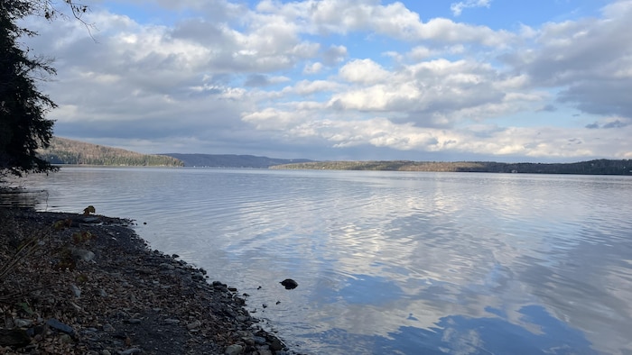 Depuis une plage rocailleuse, on voit un lac calme entouré de collines boisées.