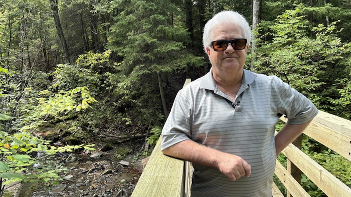 Un homme sourit, accoudé sur la garde d'une passerelle en bois qui surplombe un ruisseau dans la forêt.