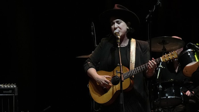 Coiffée d'un grand chapeau noir, Serena Ryder est sur scène et chante en s'accompagnant à la guitare.