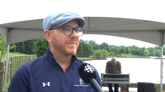 Un homme portant lunettes à monture bleue et casquette bleue accorde une entrevue près de l'eau. À l'arrière, un homme d'un certain âge est voûté au-dessus d'une grande mallette.