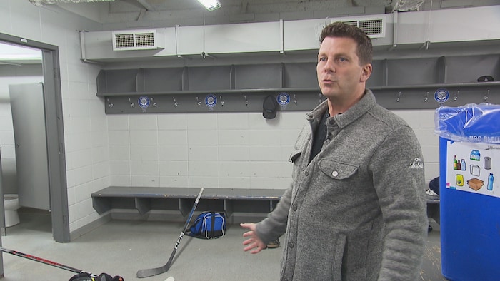 Un homme en veste grise parle dans un vestiaire de hockey. L'arrière-plan montre des bancs gris, des crochets numérotés, un bâton de hockey et un sac de sport au sol. Un bac de recyclage bleu est visible à droite.