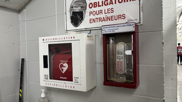 La photo montre un défibrillateur Philips dans un boîtier blanc et un téléphone d'urgence rouge fixés à un mur de blocs gris. Une affiche de sécurité et l'adresse du Centre des sports Jean-Claude Tremblay sont visibles au-dessus des équipements.
