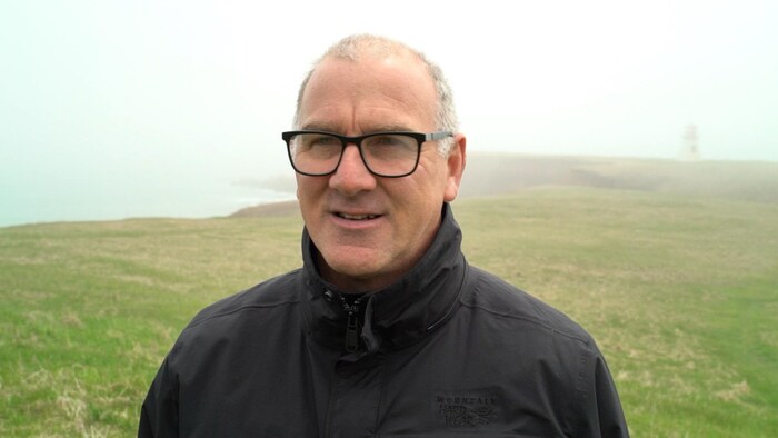 Serge Bourgeois porte des lunettes et se tient debout devant un paysage brumeux des Îles.