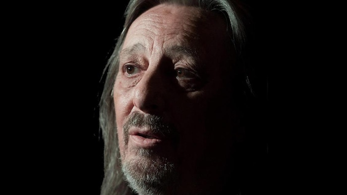 Photo du chanteur avec une moitié du visage dans l'ombre, sur fond noir. 