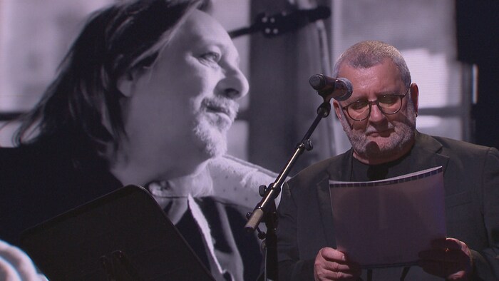 Régis Labeaume sur scène avec une photo de Serge Fiori en arrière-plan.