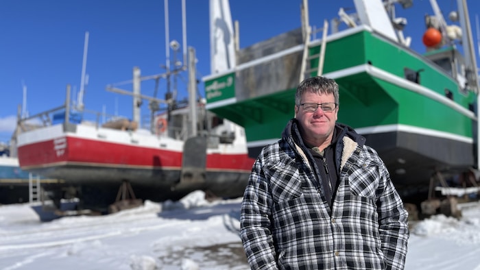 Serge Langelier, en chemise à carreaux, pose devant deux bateaux de pêche, sur un terrain enneigé. 
