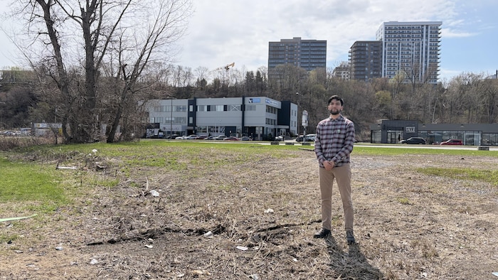 Un homme pose sur un terrain vague situé près d'un large boulevard bordé de commerces et de stationnements. Une rangée d'arbres se trouve au loin, sur une falaise surplombée de quatre grandes tours à appartements. 