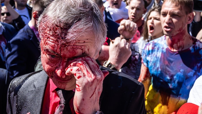 Un homme houspillé par plusieurs personnes tente de s'essuyer le visage éclaboussé d'une substance rouge.
