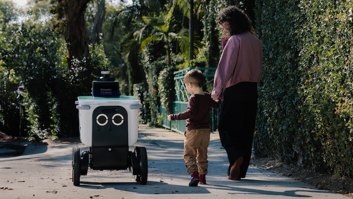 Un robot livreur dans une rue. À côté, une mère et son enfant.