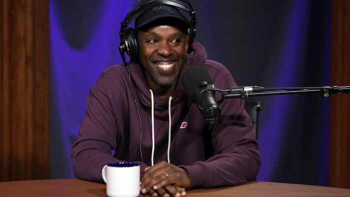 Shad sourit, assis devant un microphone dans un studio, avec un casque d’écoute sur les oreilles.