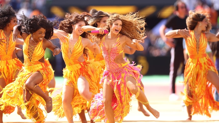 Une chanteuse entourée de danseuses danses en robes à paillettes jaunes, roses et oranges.