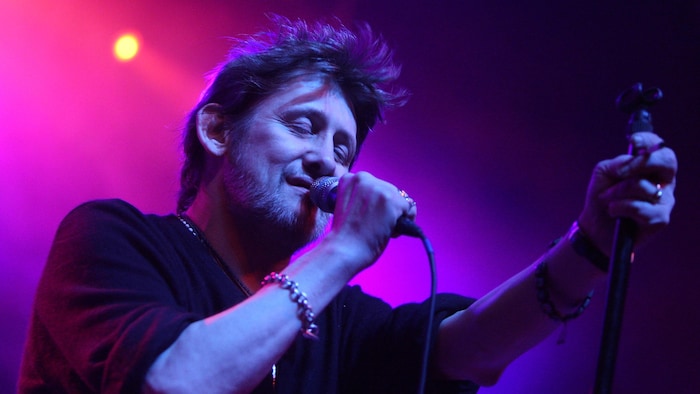 Le chanteur se produit sur scène, les yeux fermés, éclairé par une simple lumière derrière lui. 