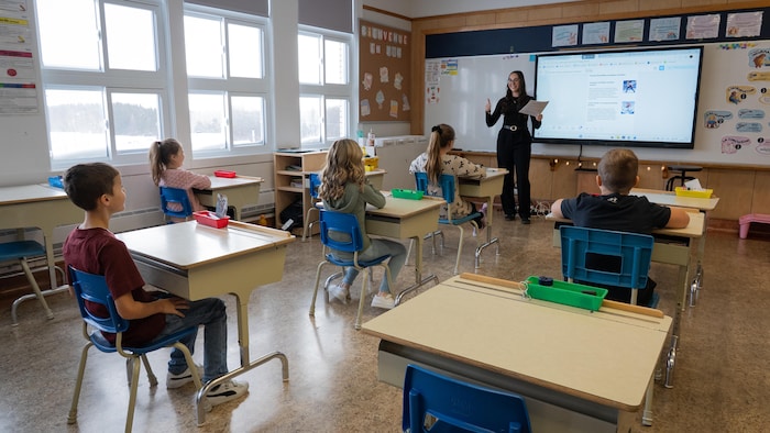 Une femme en noir à l'avant d'une classe parle à 5 élèves assis à leur pupitre devant elle.