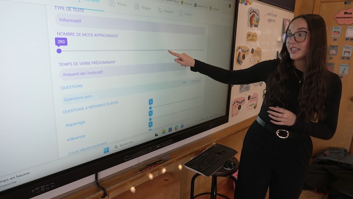 Une femme en noir pointe son doigt sur un tableau numérique de classe.