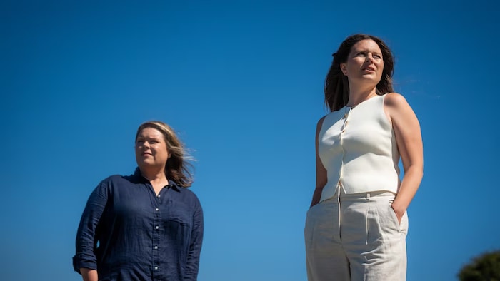 Deux femmes se tiennent debout à l’extérieur sous un ciel bleu, l’une vêtue d’une chemise bleu foncé et l’autre d’un haut blanc et d’un pantalon clair.