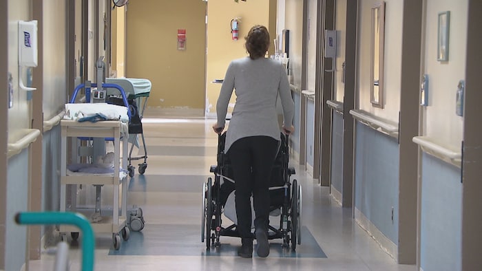 Une femme aide une personne à se déplacer en fauteuil roulant dans un couloir d'un foyer de soins.