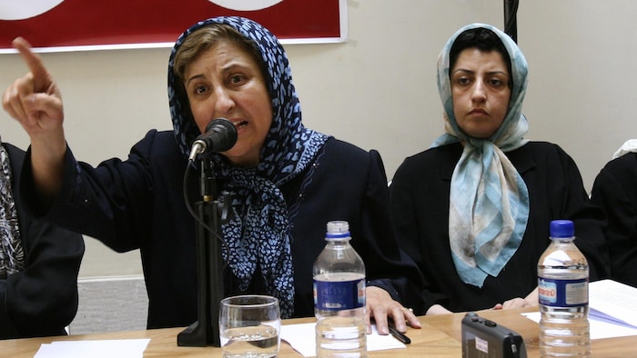 Narges Mohammadi écoute, impassible, Shirin Ebadi, qui parle au micro en arborant un air dénonciateur, l'index levé.