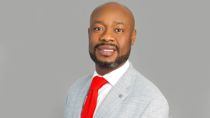 Shola Agboola porte une chemise blanche, une cravate rouge et un veston gris. Il est photographié devant un fond gris. (2025)