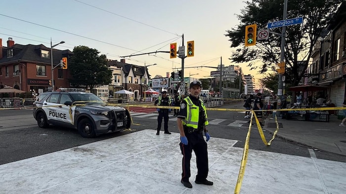 Un policier regarde vers l’appareil photo. Il se tient près d’une zone délimitée par un ruban de sécurité jaune, à l’angle des avenues Arlington et St. Clair Ouest à Toronto. En arrière-plan, on aperçoit des tentes de festival et d’autres policiers assurant la sécurité du périmètre.