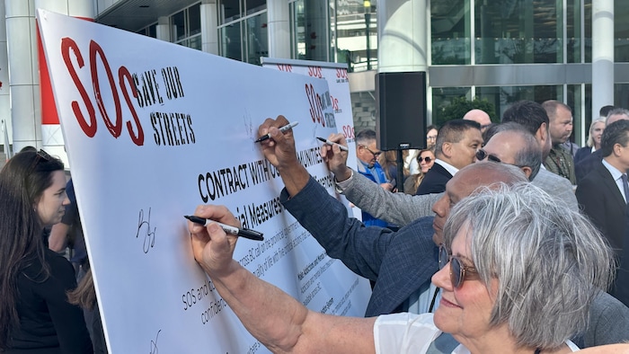 Plusieurs personnes signent simultanément au marqueur un grand panneau blanc portant le logo SOS Save Our Streets, devant le Centre des congrès de Vancouver.