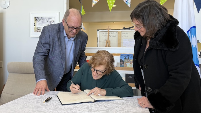 Hugues Lafrance, maire suppléant de la municipalité des Îles-de-la-Madeleine, regarde Lucie Cormier signer le livre d'or aux côtés de sa fille, Laurie Lapierre.